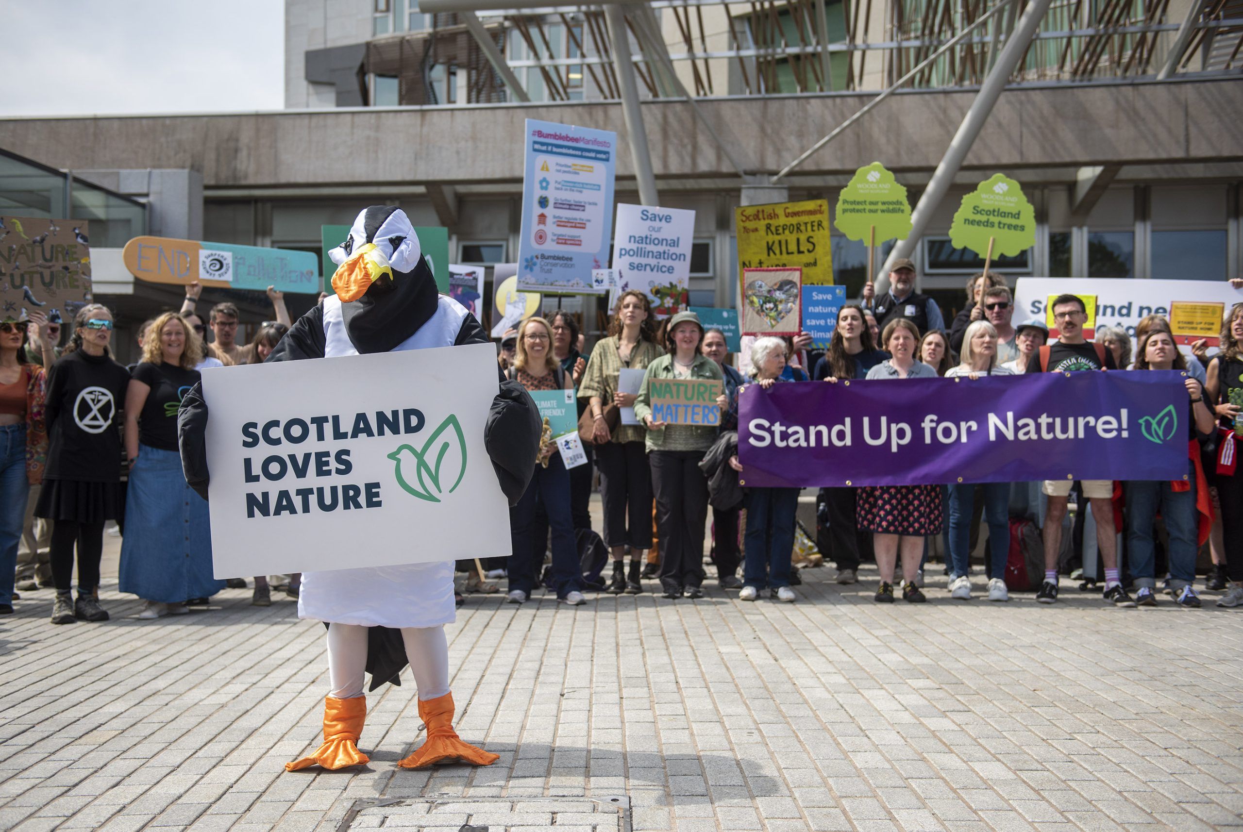 A group of people standing outside the Scottish parliament holding colourful signs which say things like Scotland Loves Nature and Stand Up for Nature, with a person in a puffin costume at the front.