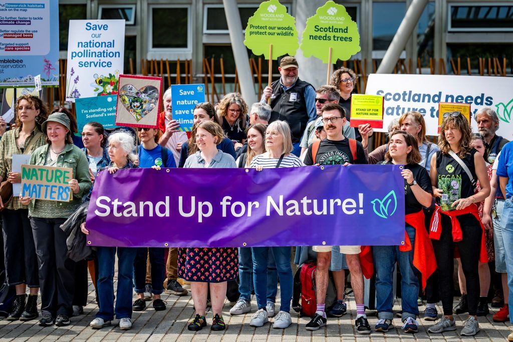 People with signs and banners standing outside the Scottish parliament building. A purple banner in the foreground reads, 'Stand Up for Nature!'. Other signs read, 'Nature matters', 'Save our national pollination service', 'Protect our woods, protect out wildlife', and 'Scotland needs more trees'.
