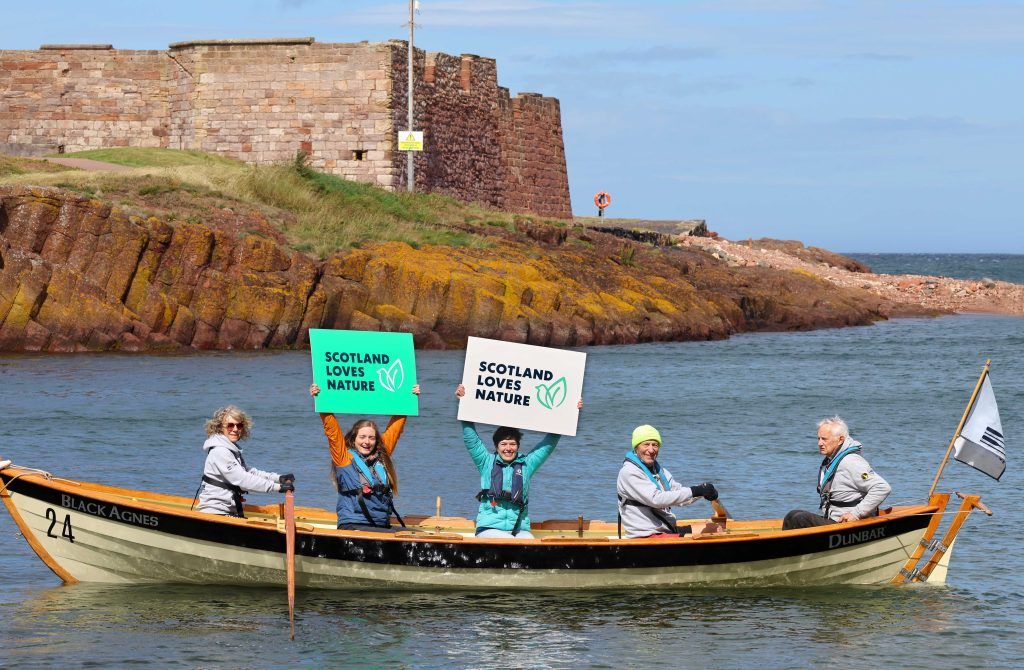Five people in a rowing boat on the sea, two of whom are holding signs reading 'Scotland Loves Nature', and some rocks and an old stone structure in the background